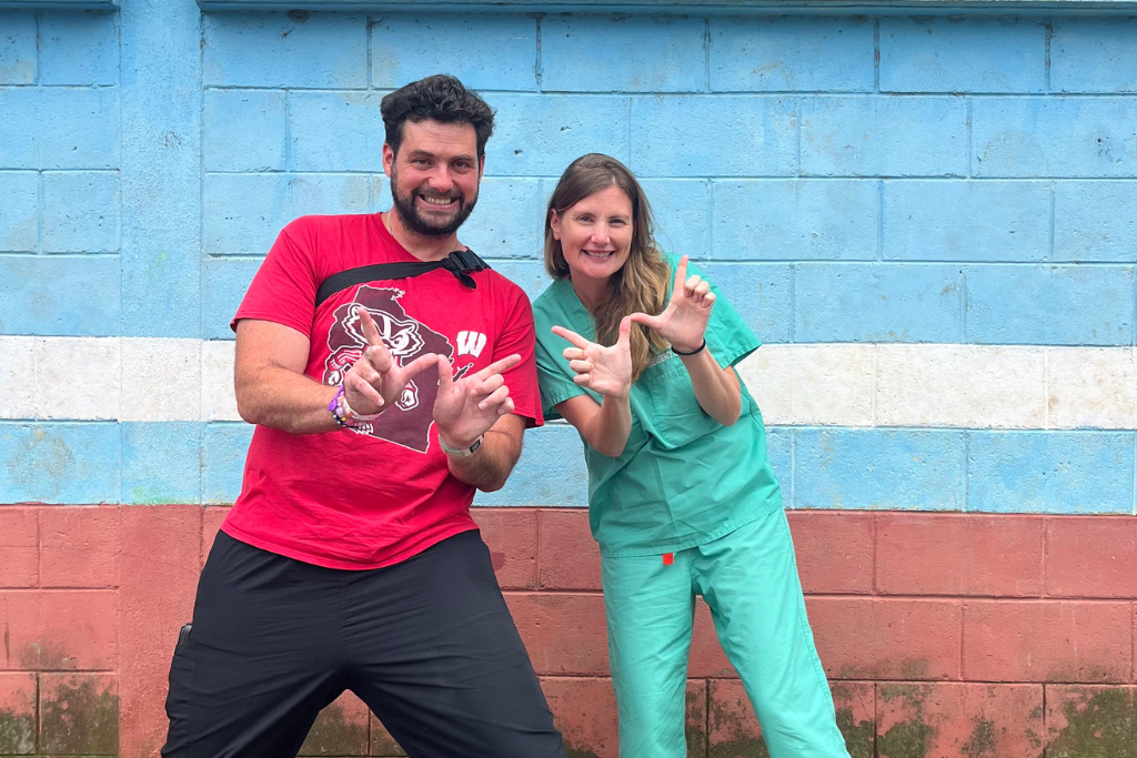 Two people posing playfully with raised hands in front of a blue wall, one wearing a red T-shirt and the other green medical scrubs.